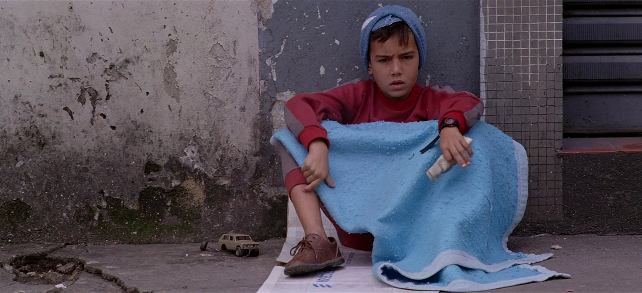 Baraka (1992) movie still: medium — A young boy sits on a paved surface against a weathered, peeling exterior wall. He is wrapped in a light blue…