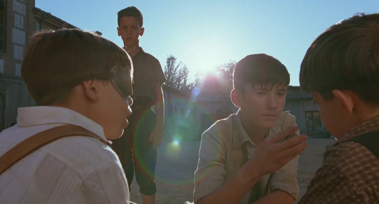 The Devils Backbone (2001) movie still: medium — A stone courtyard during the day with sun glare appearing behind the group. Four young boys stand…