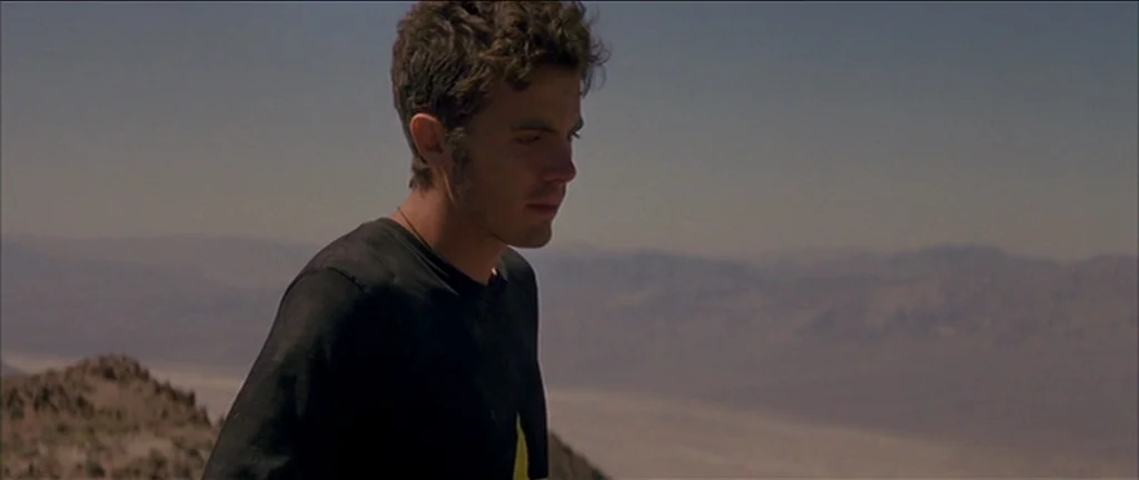 Gerry (2002) movie still: medium — A man stands atop a rocky terrain overlooking a vast, dry desert landscape. The subject looks down while the…