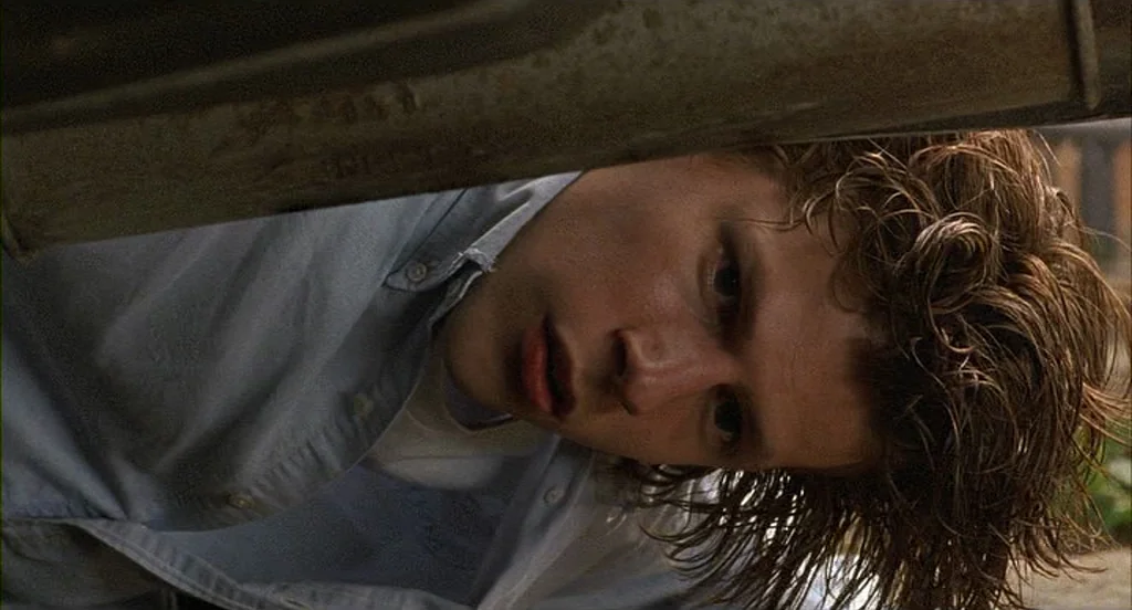 The Squid and the Whale (2005) movie still: closeup — A man lies on the ground peering from beneath the chassis of an automobile. The metal frame of…