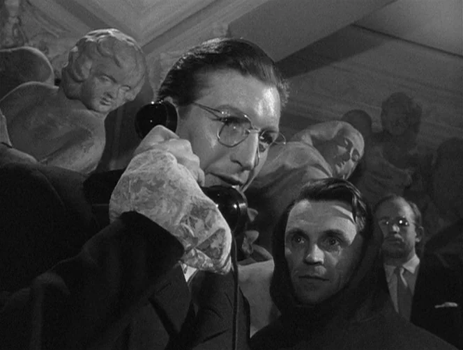 Mr. Arkadin (1955) movie still: medium — A dark, crowded interior space filled with classical marble sculptures of figures. A man in glasses uses a…