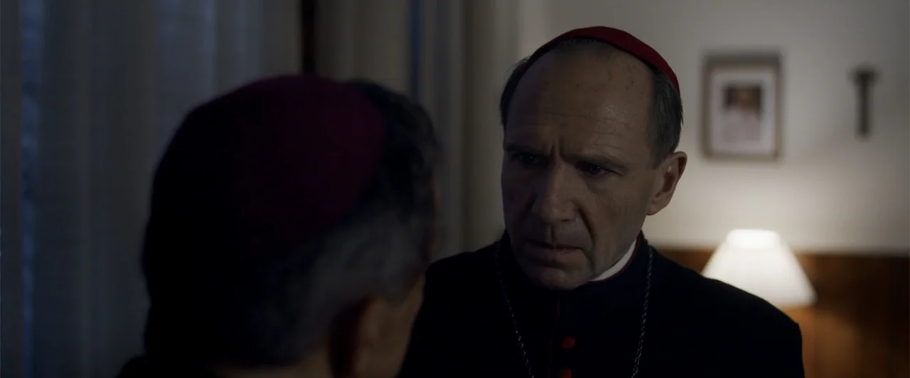 Conclave (2024) movie still: closeup — A dimly lit room with a desk lamp and a framed picture on the wall. A cardinal in black robes leans in to…