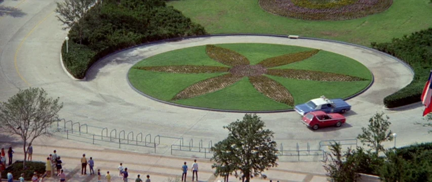 Brewster McCloud (1970) movie still: establishing — A wide aerial view shows a circular driveway surrounding a circular lawn with patterns. Two…
