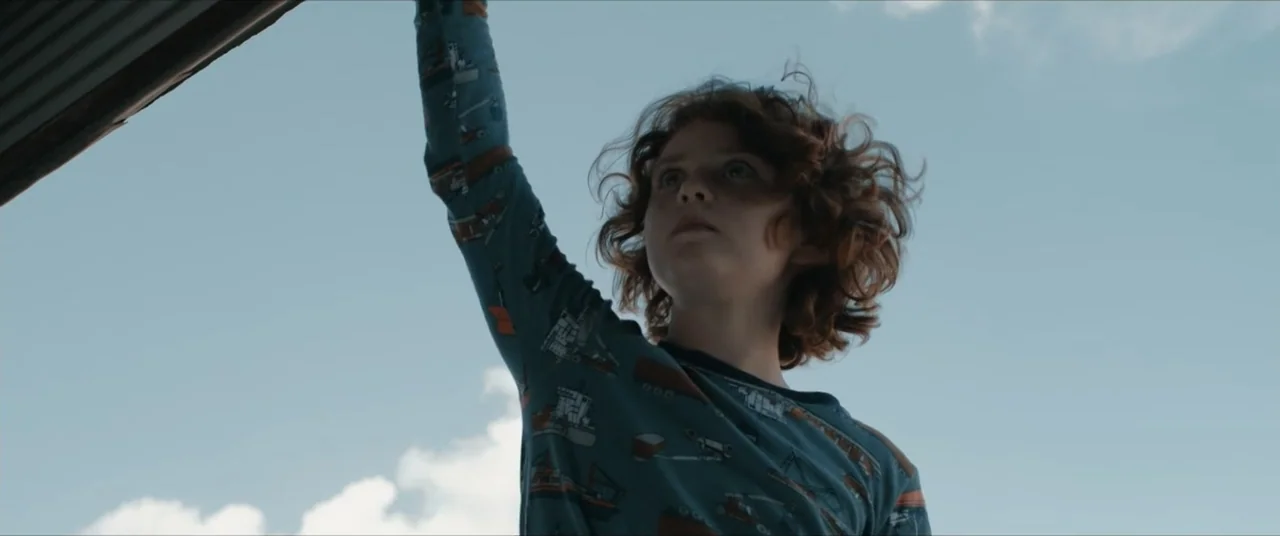 Possessions (2024) movie still: medium — An open exterior space with a clear blue sky and thin white clouds above. A young boy in a long-sleeved…