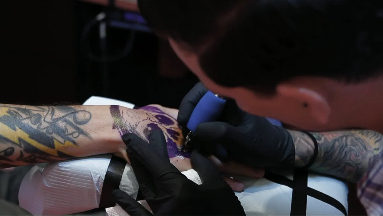 Control (2022) movie still: closeup — A close-up view shows a hand resting on a white sanitary towel. A tattoo artist wearing black gloves holds a…