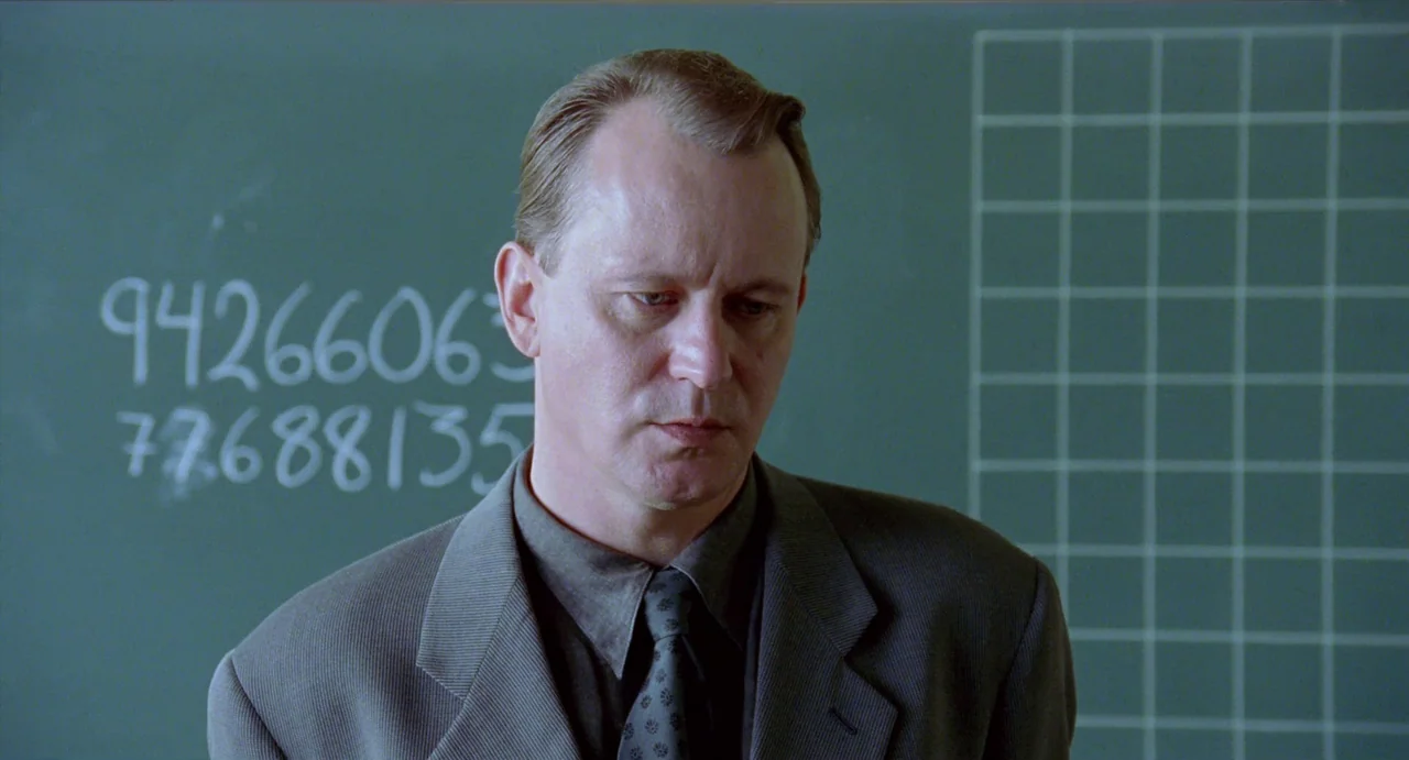 Insomnia (1997) movie still: medium — A man stands before a dark green chalkboard with grid markings and white numbers. He is positioned slightly to…