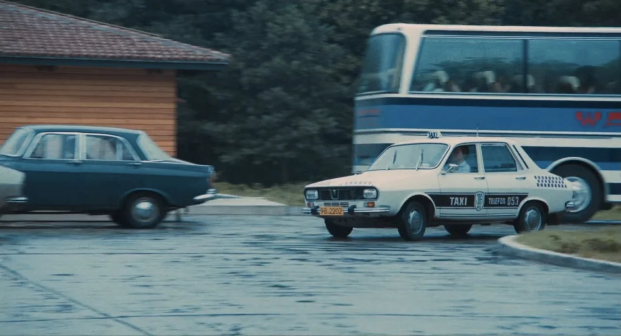 Do Not Expect Too Much from the End of the World (2023) movie still: wide — A wet asphalt parking lot features several vintage vehicles moving past…