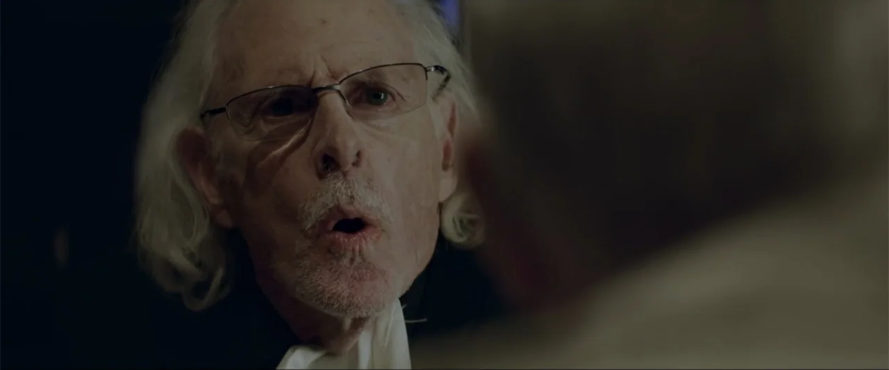 Gender Under Attack (2018) movie still: closeup — A dimly lit interior space features an elderly man in the foreground. He is engaged in a…