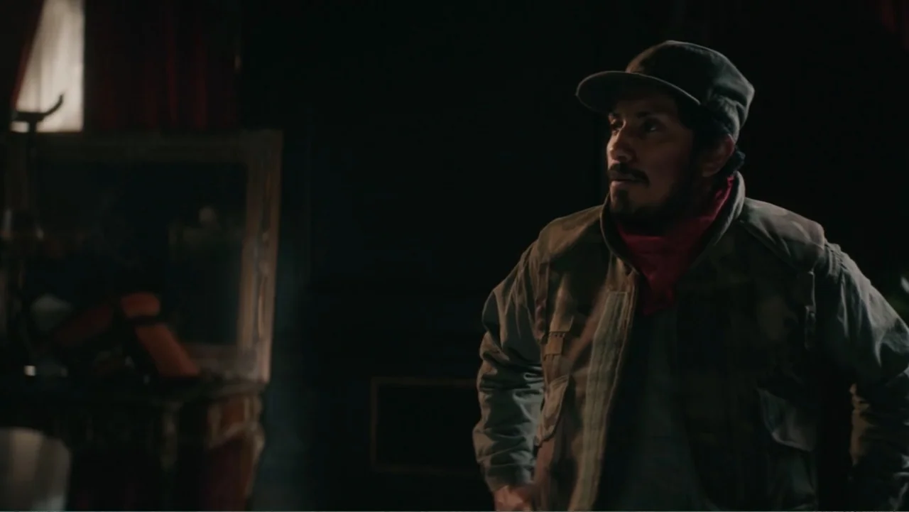 Bel Canto (2018) movie still: medium — A dimly lit room contains a mirror and draped fabrics in the shadows. A man wearing a camouflage jacket and…