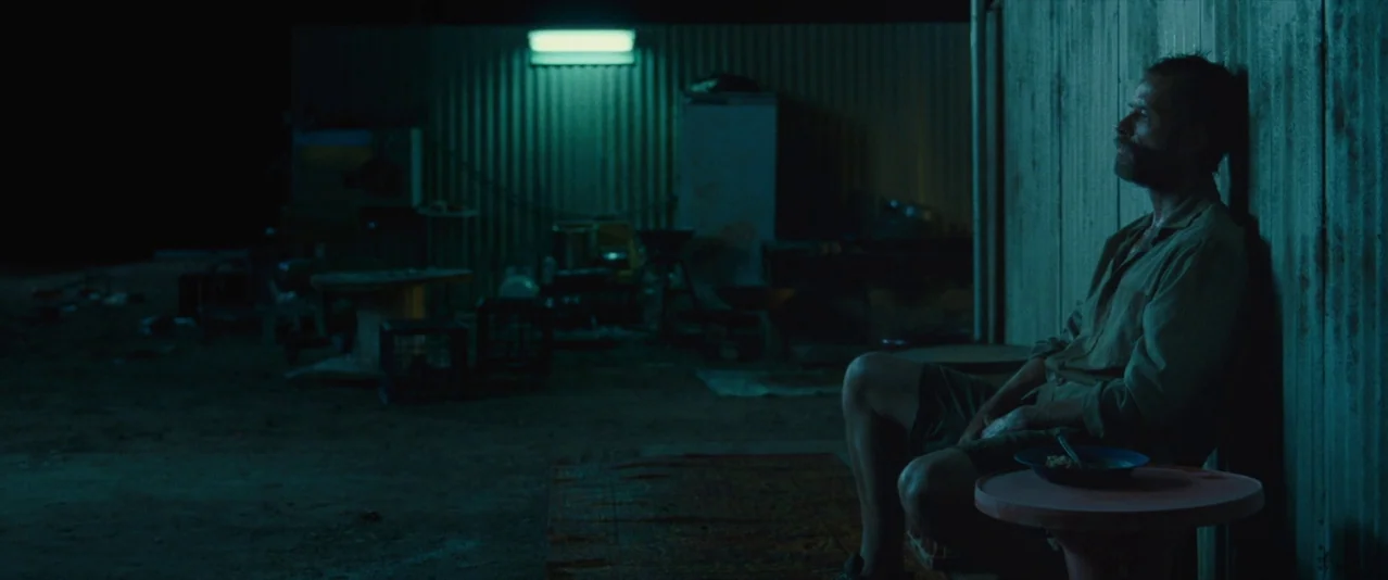 The Rover (2014) movie still: medium — A man sits against a rusted corrugated metal wall in a dark industrial yard. A single bowl of food rests on a…
