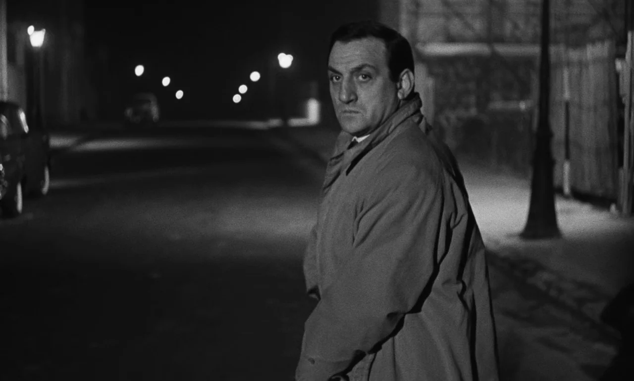 Witness In The City (1959) movie still: medium — A dark, empty city street at night illuminated by distant streetlights. A man in a trench coat…