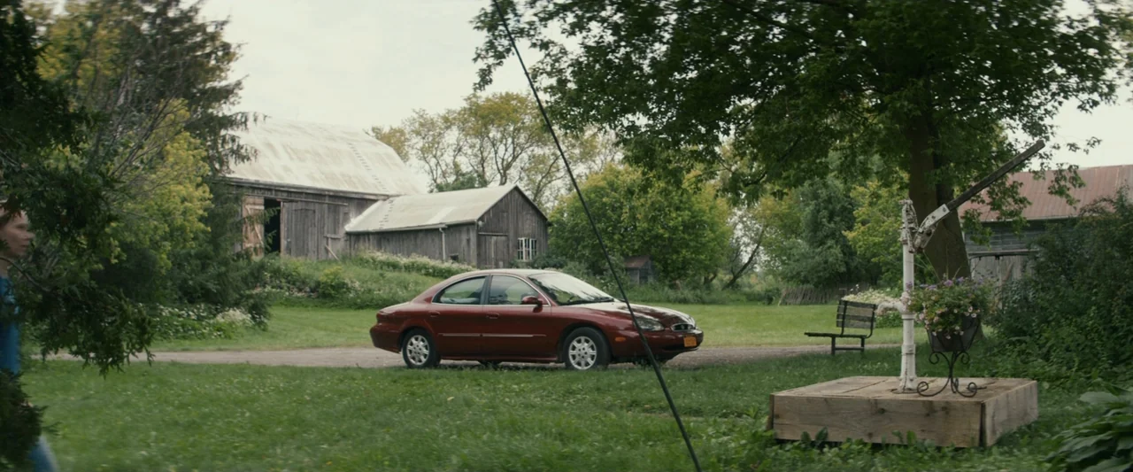 Strange but True (2019) movie still: wide — A rural property features a red sedan parked on a gravel path near wooden barns. A person walks through…