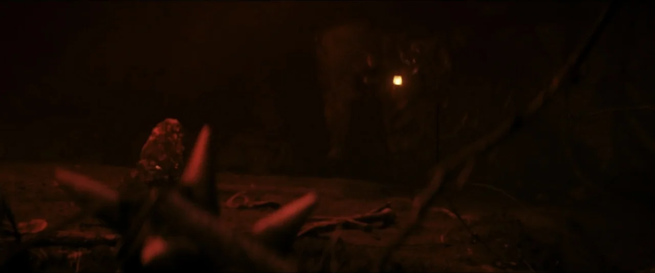 It Feeds (2025) movie still: wide — A dark subterranean cavern with rocky textures and scattered debris on the floor. A faint, distant light source…