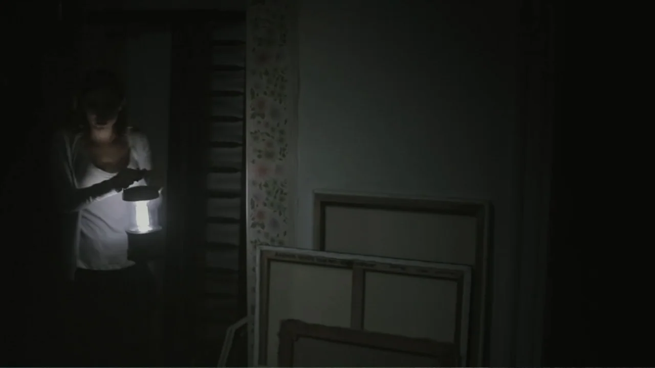 Silent House (2011) movie still: medium — A dark, dimly lit room contains stacked blank canvases and patterned floral wallpaper. A young woman…