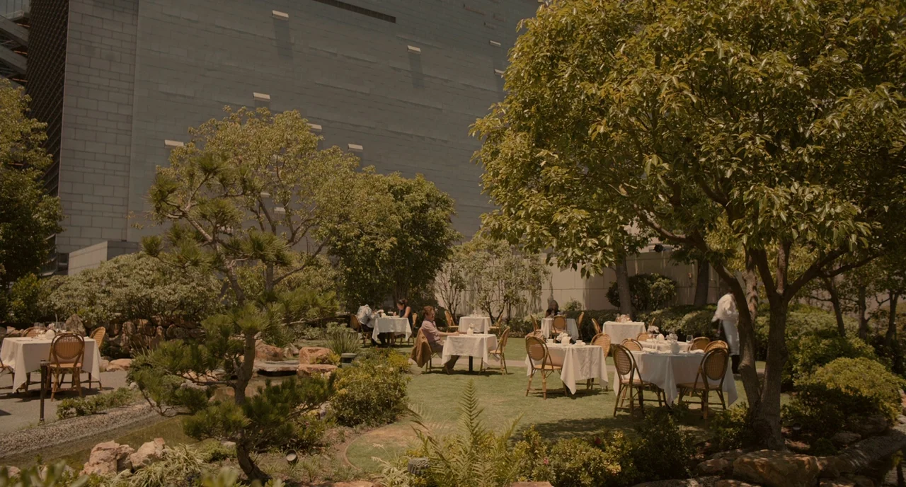 Her (2013) movie still: wide — A landscaped outdoor terrace features multiple tables with white cloths set among mature trees. A man sits at a table…