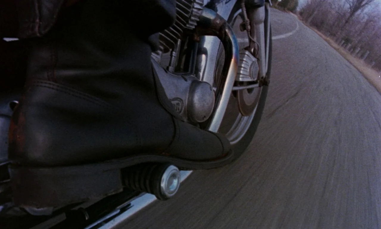Rabid (1977) movie still: closeup — A close perspective shows a black boot resting on a motorcycle foot peg. The motorcycle moves along a grey paved…
