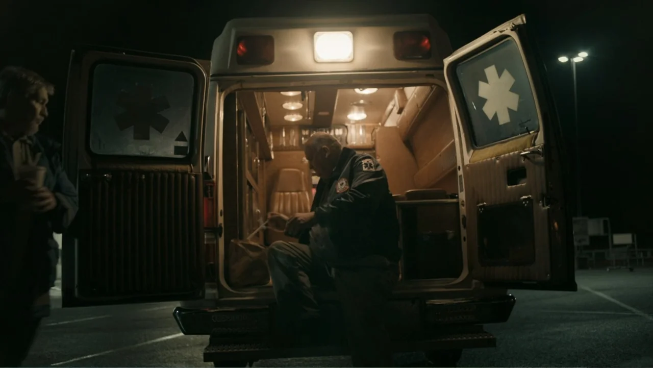 Alabama Snake (2020) movie still: wide — A nighttime parking lot contains an open EMS vehicle with its rear doors swung wide. A man in an EMS jacket…