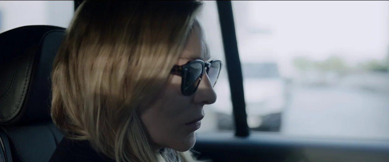 Tár (2022) movie still: closeup — The interior of a vehicle passenger cabin shows the side profile of a woman. A blonde woman in dark sunglasses…