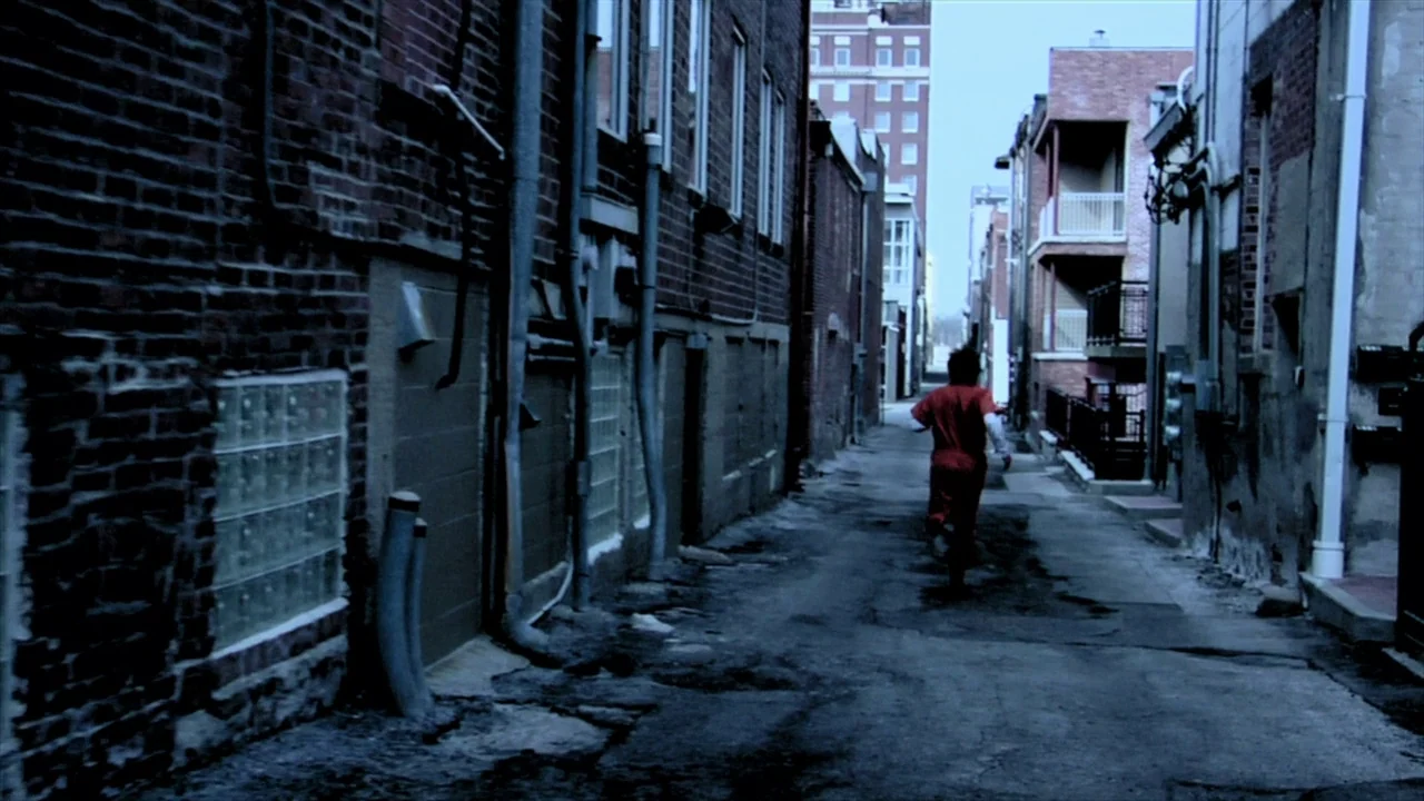 A Horrible Way To Die (2010) movie still: wide — A narrow city alleyway is flanked by red brick walls with vertical drainage pipes. A person in an…