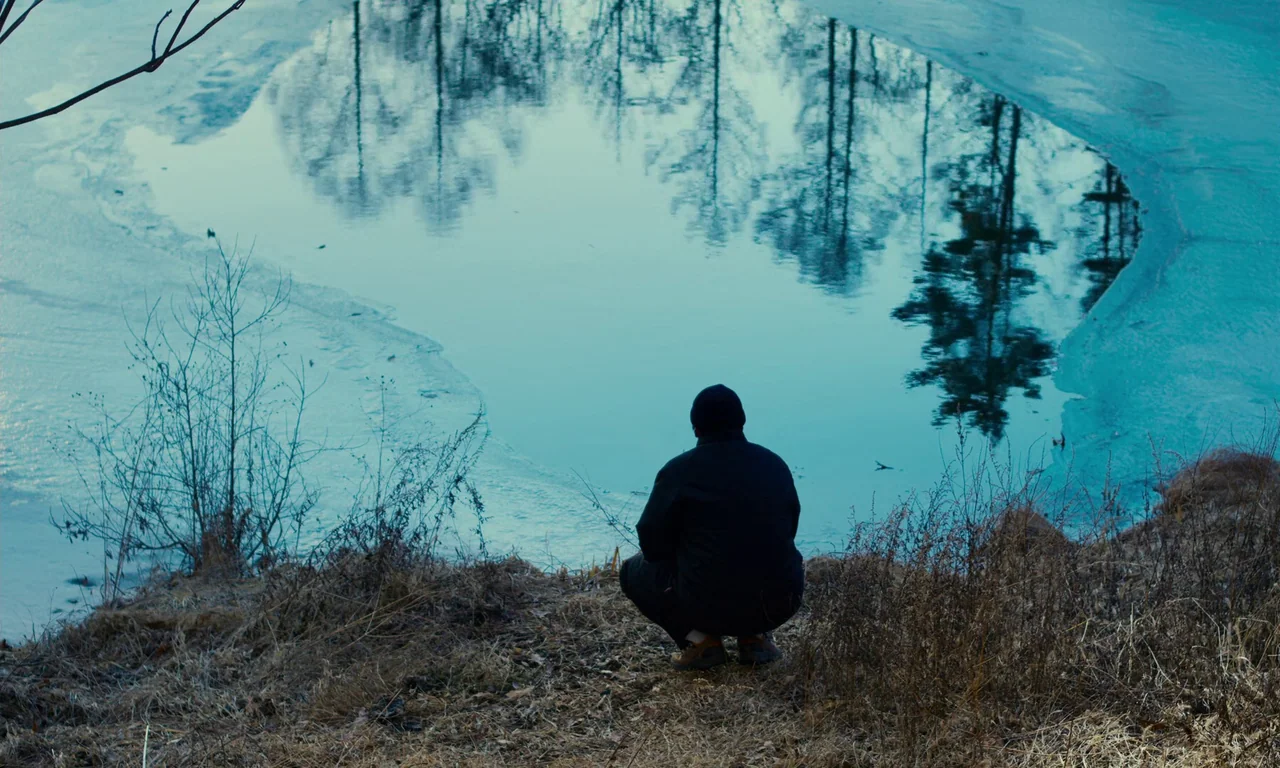 Evil Does Not Exist (2023) movie still: wide — A man crouches on the dry, dormant grass at the edge of a partially frozen lake. Tall trees reflect…