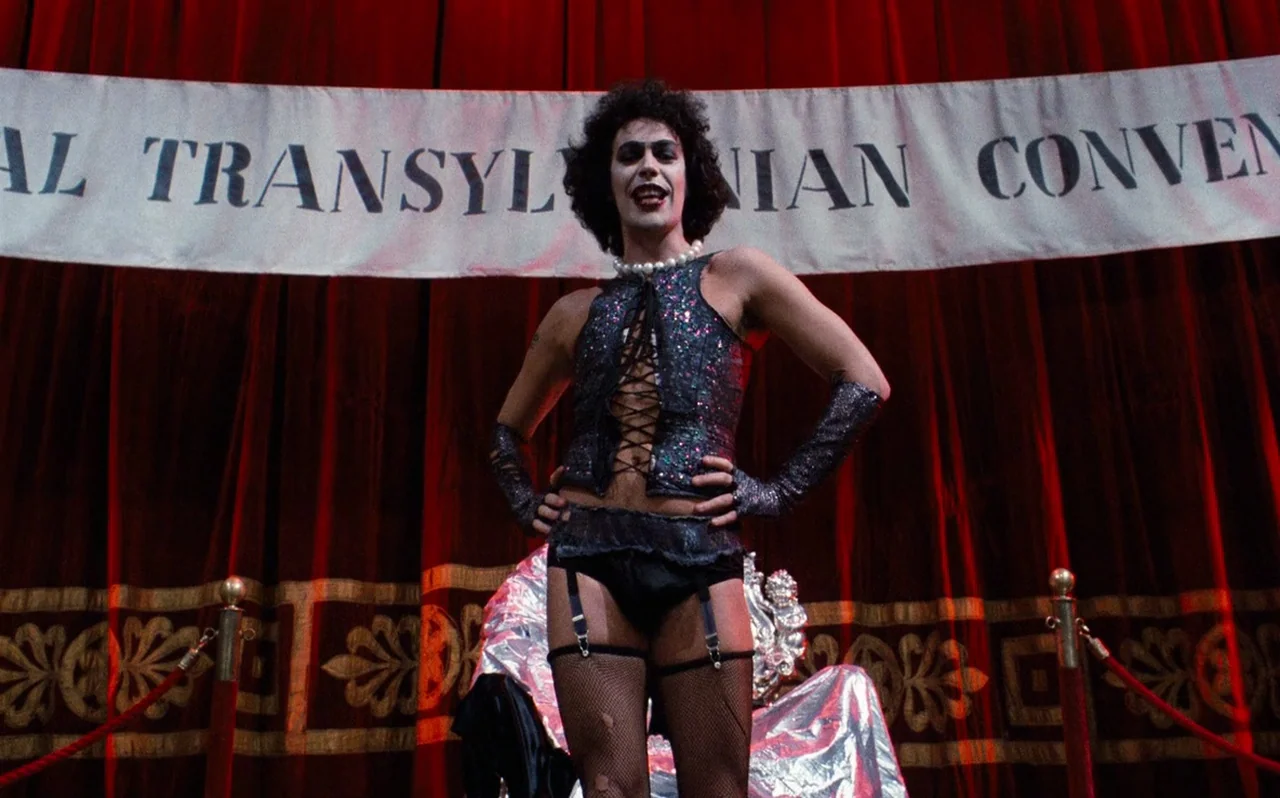 The Rocky Horror Picture Show (1975) movie still: fullbody — A theatrical stage features heavy red velvet curtains and a draped banner overhead. A…