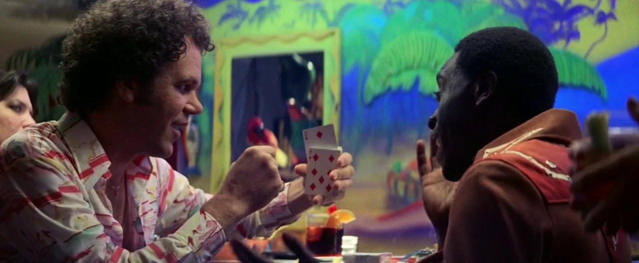 Boogie Nights movie still: medium — A bar interior features a vibrant hand-painted tropical mural with palm trees and leaves. A man with curly hair…
