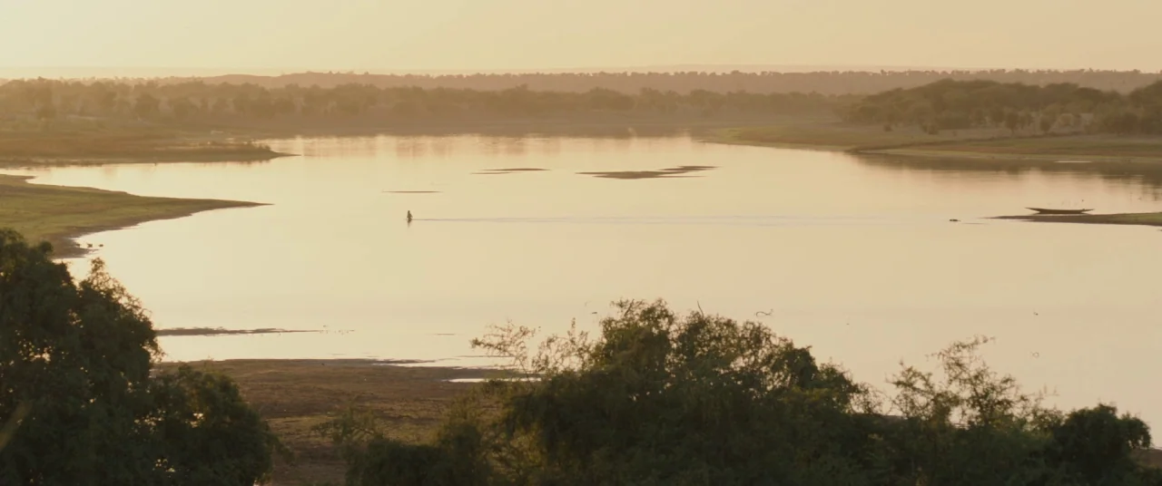 Timbuktu (2014) movie still: wide — A wide river winds through a flat, sparsely vegetated landscape under a hazy sky. A single person is wading…
