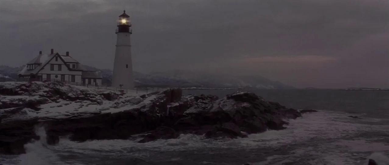 Snow Falling on Cedars (1999) movie still: establishing — A tall white lighthouse stands next to a wooden keeper house on a rocky cliff. Snow covers…