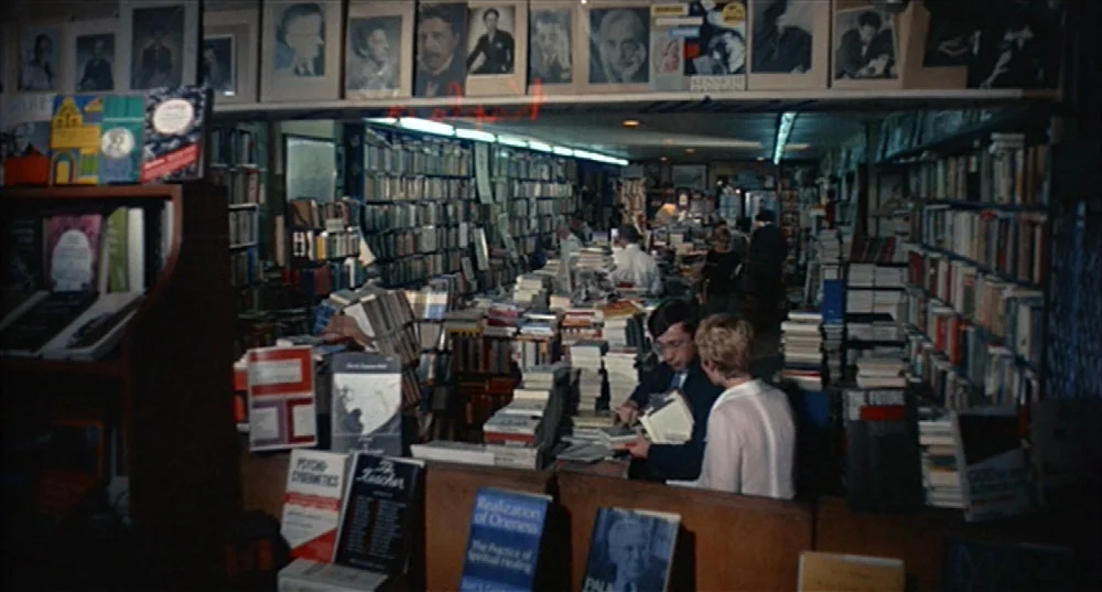 Rosemary’s Baby (1968) movie still: wide — A dense bookstore interior is filled with floor-to-ceiling shelves and stacks of paperbacks. A couple…