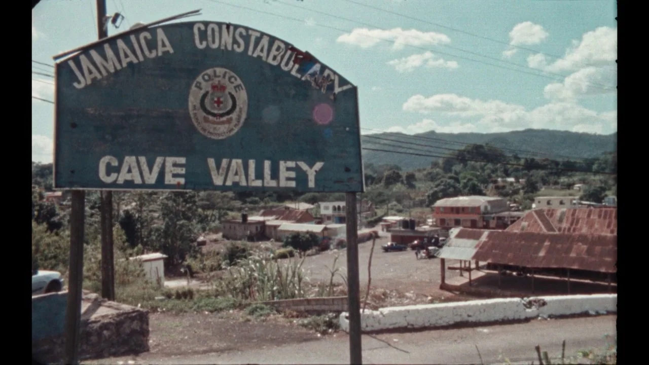 Black Mother (2018) movie still: establishing — A weather-worn sign for the Jamaica Constabulary stands overlooking a cluster of small buildings.…