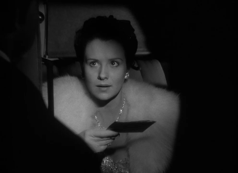 Citizen Kane (1941) movie still: medium — A dark car interior with a woman sitting in the passenger seat. She holds a small rectangular card toward…