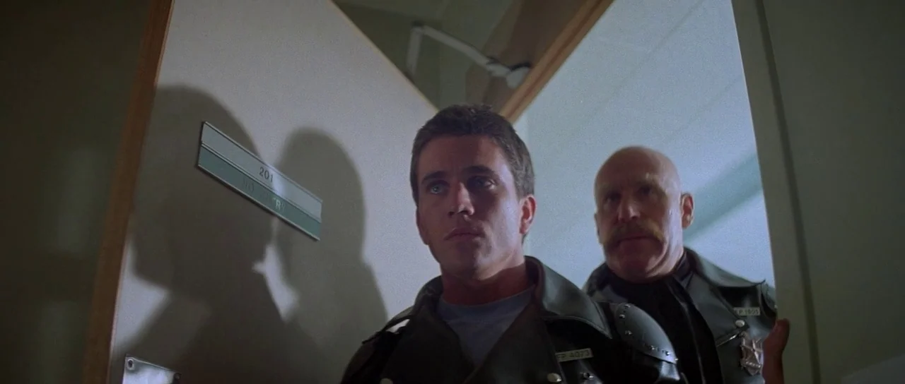 Mad Max (1979) movie still: medium — An interior doorway with a sign marked 201 opens into a sterile hallway. Two men in leather jackets move…