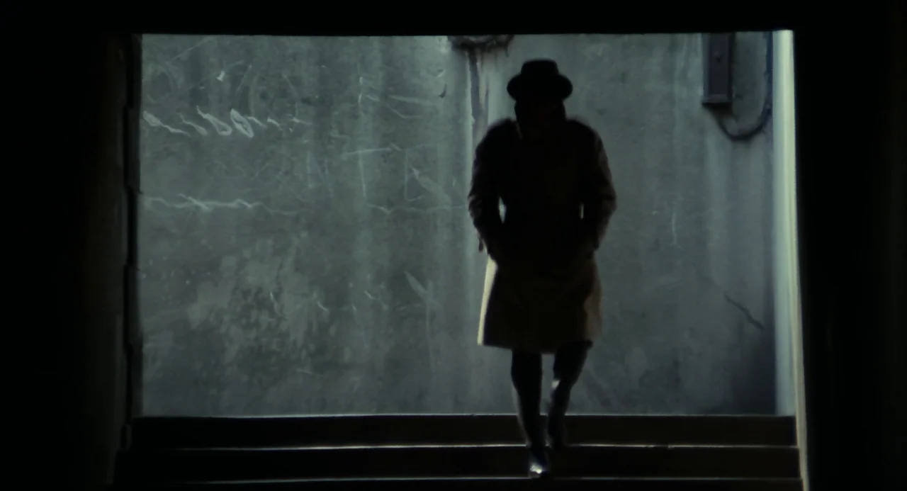Le Samouraï (1967) movie still: fullbody — An outdoor concrete stairwell features weathered walls with faint markings and heavy shadows. A figure in…