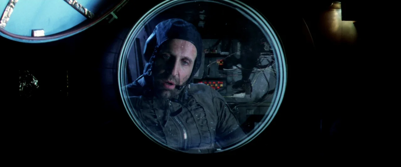 Armageddon (1998) movie still: closeup — A man looks through a circular thick glass window into a dark technical compartment. Exposed wires and…
