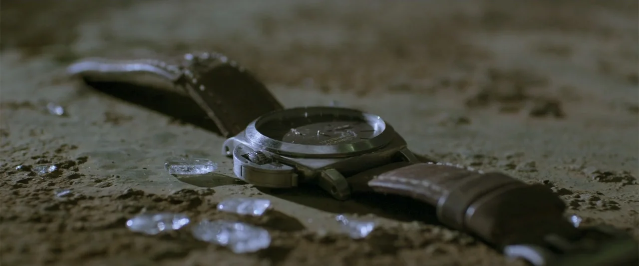 Skiptrace (2016) movie still: closeup — A wristwatch with a brown leather strap lies on a rough, textured concrete surface. Several sharp shards of…