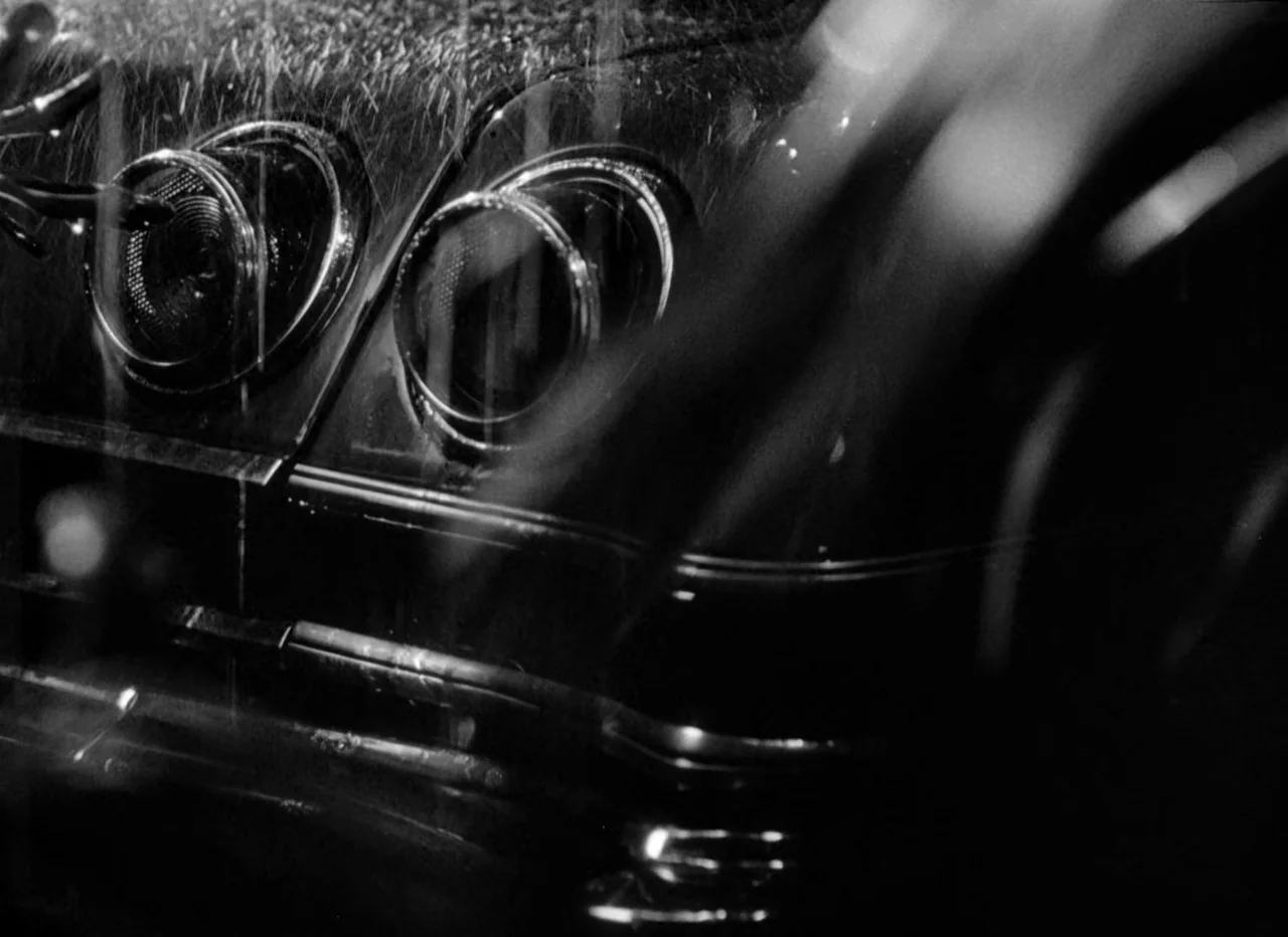 Singapore Sling (1990) movie still: closeup — The rear lights and fender of a classic automobile are visible in the rain. Water droplets streak…