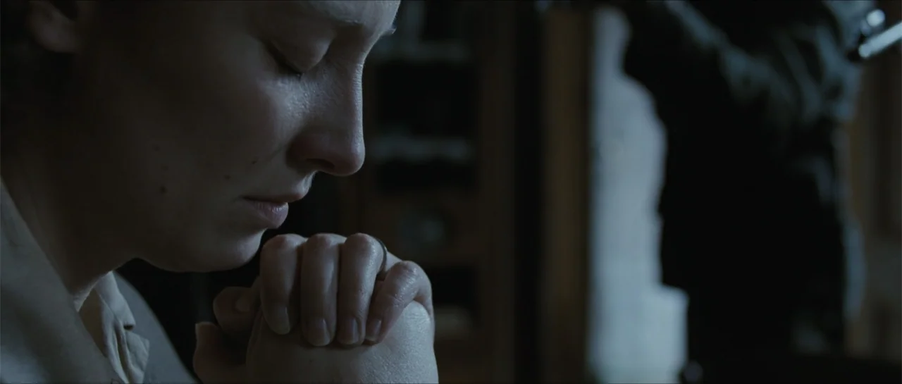 Suite Française (2015) movie still: closeup — An interior space contains a person in soft focus and low-light conditions. A woman with short hair…