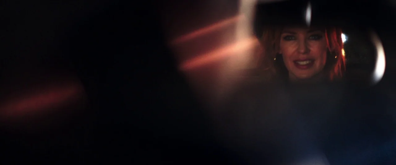 20,000 Days on Earth (2014) movie still: closeup — A dark vehicle interior with a view of a woman seen through a mirror. The woman sits in the…