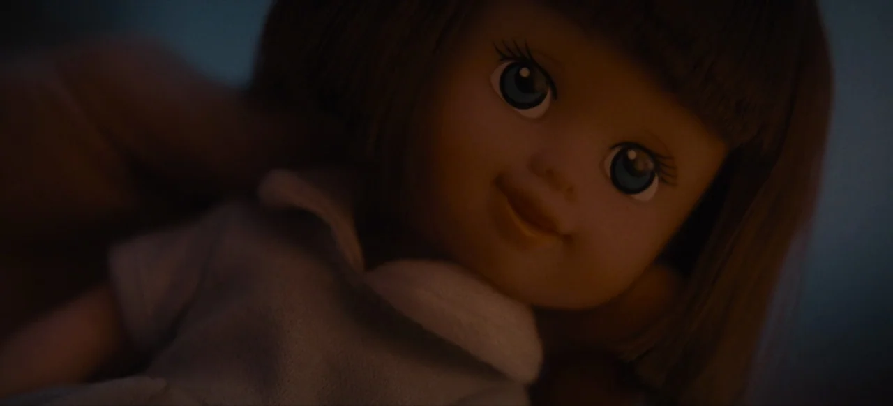 Housewife (2017) movie still: closeup — A close-up view of a plastic doll's face in low light. The doll looks toward the camera with painted blue…