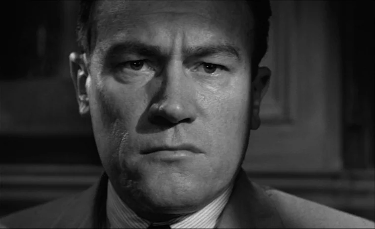 12 Angry Men (1957) movie still: closeup — A man stands in an interior space with dark, nondescript background paneling. He is looking straight…