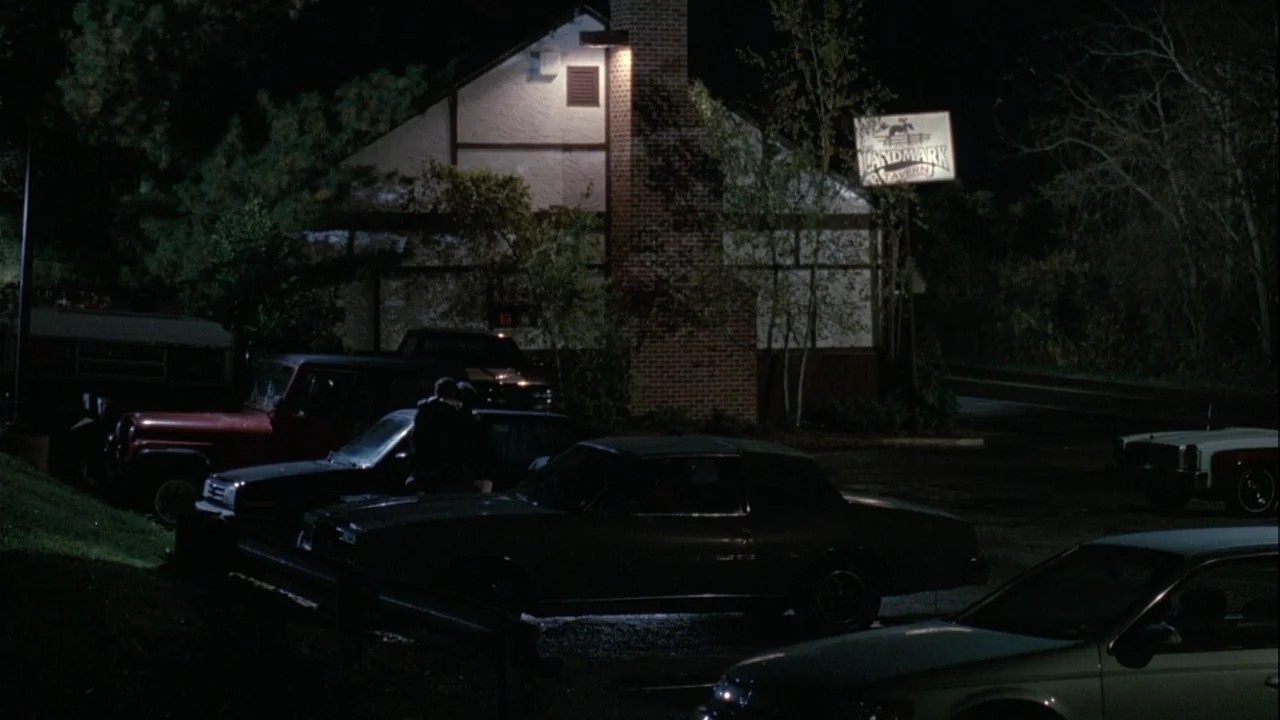 Boys (2021) movie still: wide — A nighttime parking lot features a tavern building with timber-framed walls and a lit sign. Two people stand close…