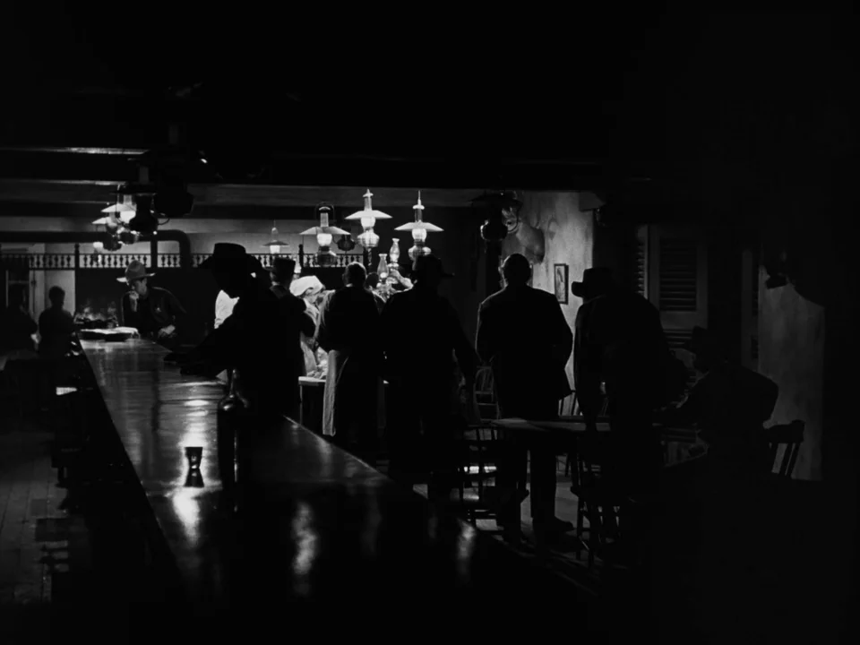 My Darling Clementine (1946) movie still: wide — A dimly lit western saloon features a long polished wooden bar and hanging lamps. Several men in…