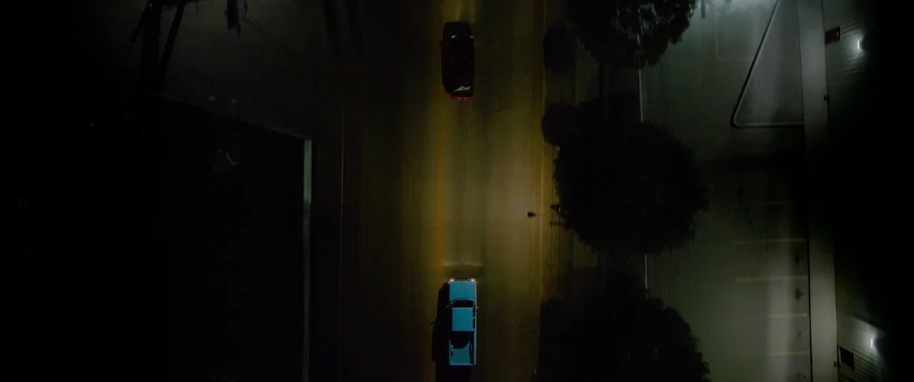 Love Lies Bleeding (2024) movie still: wide — A dark road at night is captured from an overhead top-down perspective. Two cars move past each other…