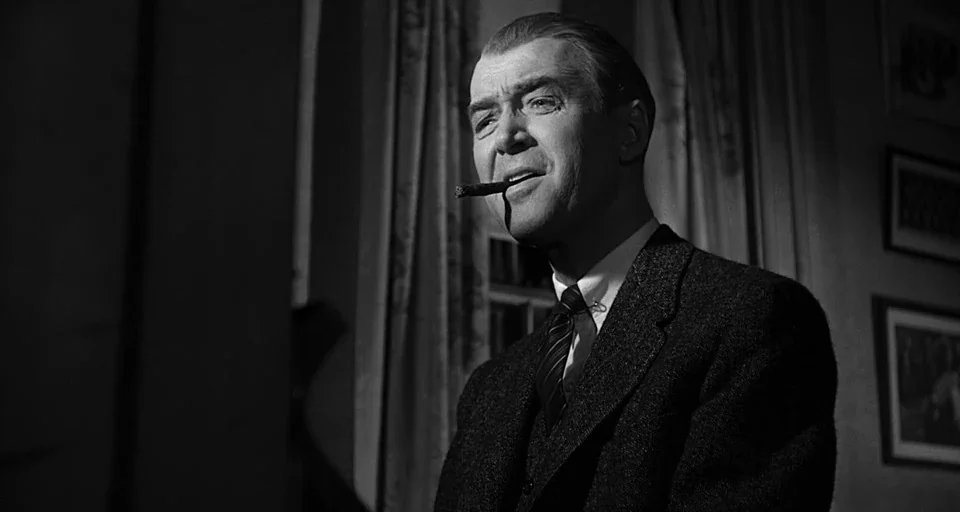 Anatomy of a Murder (1959) movie still: medium — A dimly lit room features a man standing in front of patterned fabric curtains. He wears a tweed…