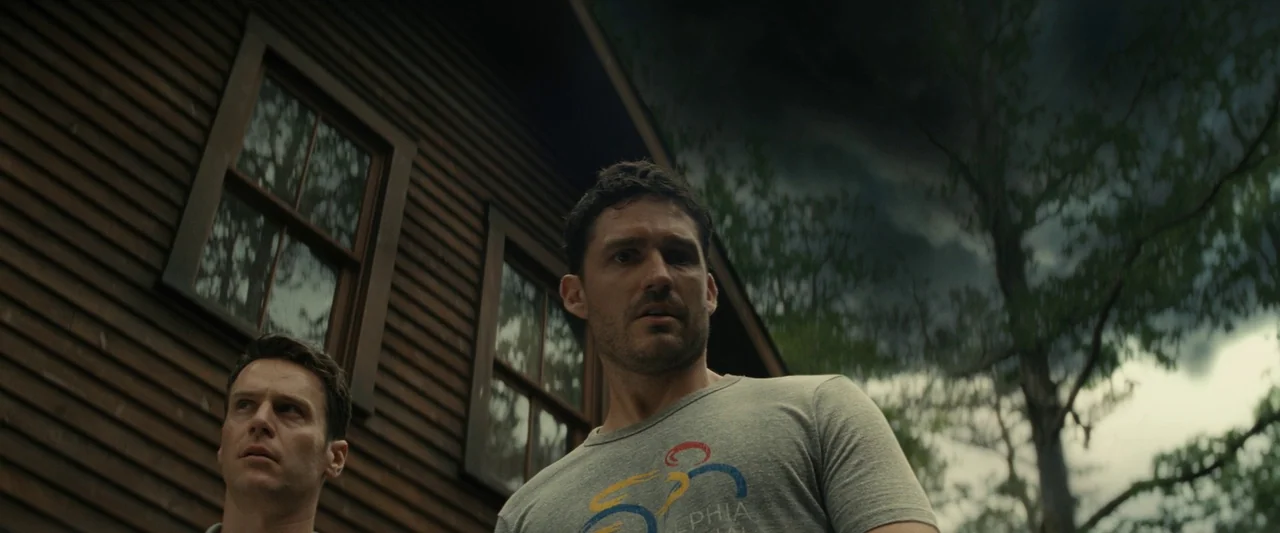 Knock at the Cabin (2023) movie still: medium — A dark, wooden cabin structure stands against a clouded sky with mature trees nearby. Two men stand…