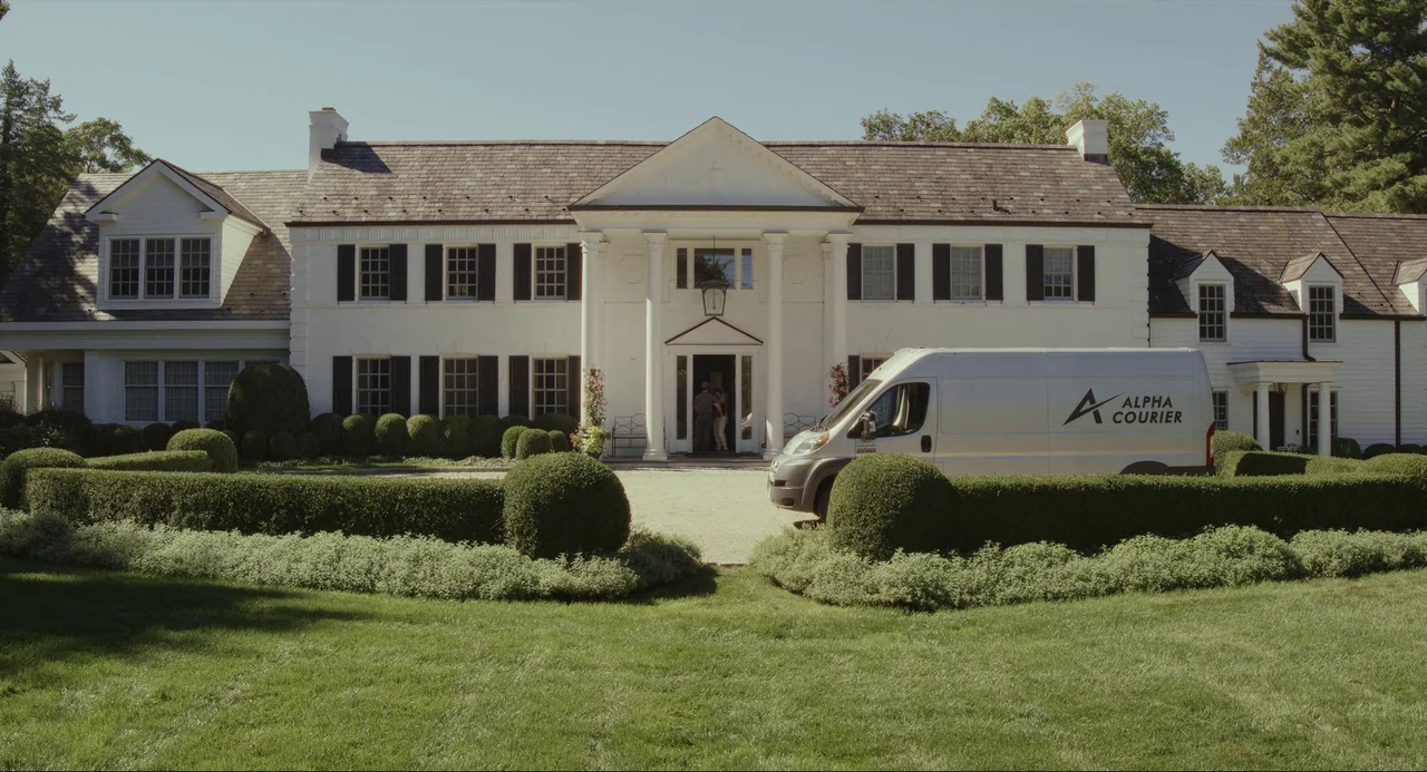 Glass Onion (2022) movie still: wide — A large white two-story mansion with black shutters sits behind a manicured green lawn. A silver delivery van…