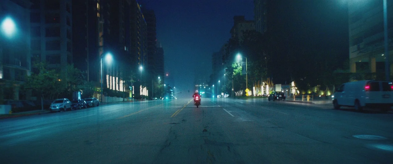 Gemini (2017) movie still: wide — A wide city street at night is framed by apartment buildings and parked cars. A lone motorcycle travels away from…