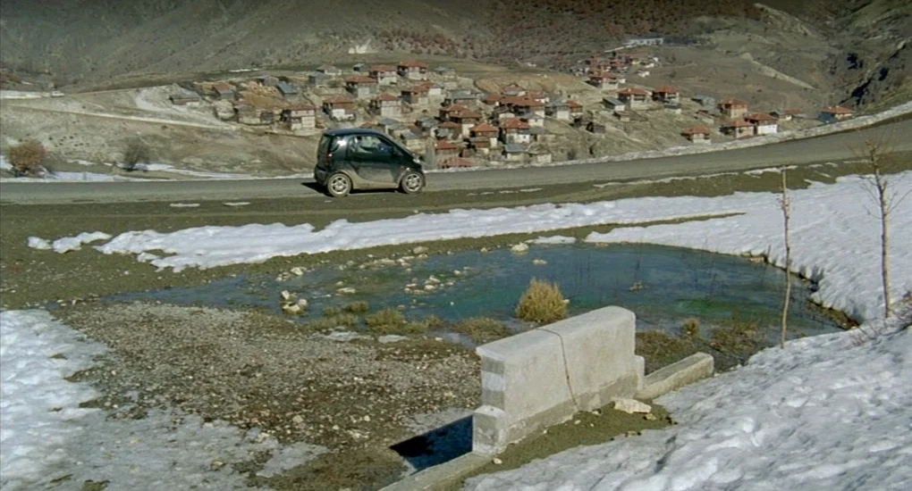 Uzak (2002) movie still: wide — A small black car sits on a paved road overlooking a village nestled in mountains. A patch of melting snow surrounds…