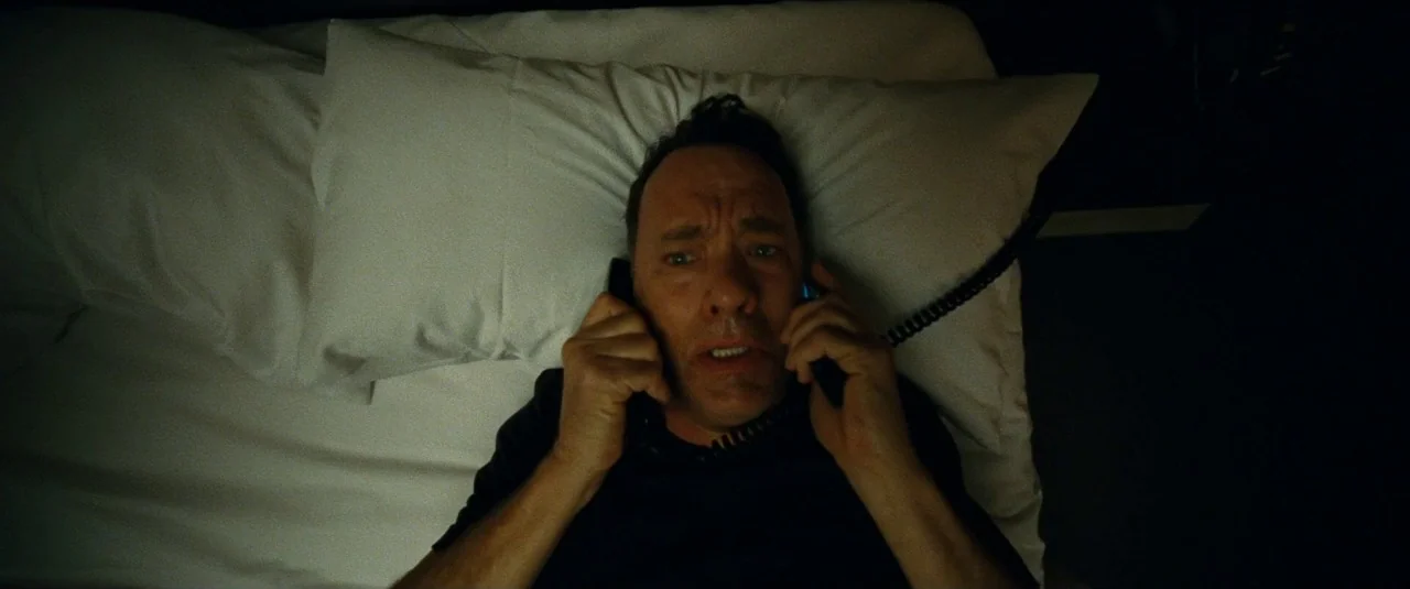 A Hologram for the King movie still: closeup — A man lies on a white pillow in a dark, dimly lit environment. He holds a black telephone handset to…