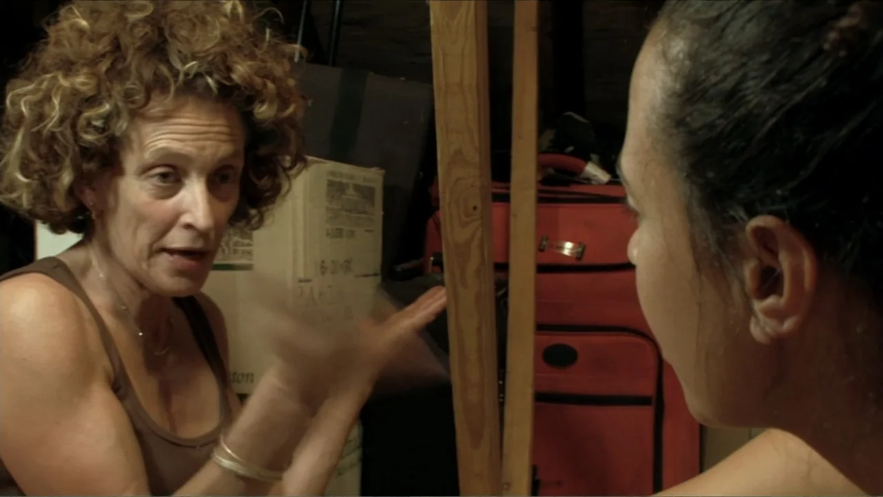 Little White Lie (2008) movie still: medium — A cluttered storage space filled with stacked cardboard boxes and red luggage bags. A woman with curly…